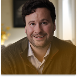 Man in a white button-down and brown sweater smiling